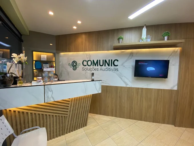 Recepção da Comunic Soluções Auditivas: balcão de atendimento em madeira e mármore, com o logotipo da marca na parede